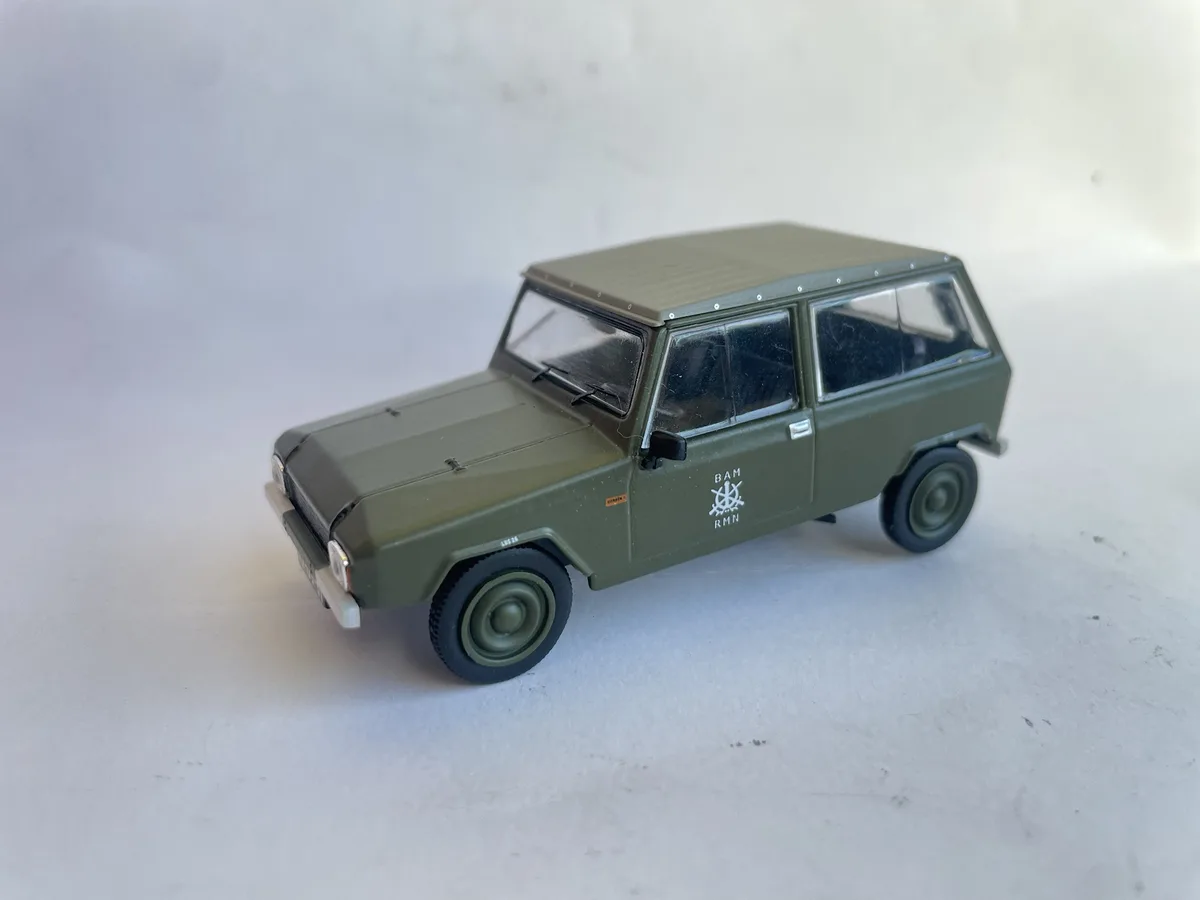 A green toy model of a vintage military-style station wagon is photographed against a plain white background. The side of…