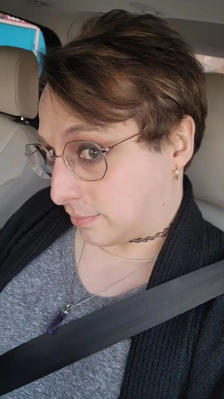 A person with short brown hair, glasses, and multiple necklaces sits in a car wearing a seatbelt that crosses over their…