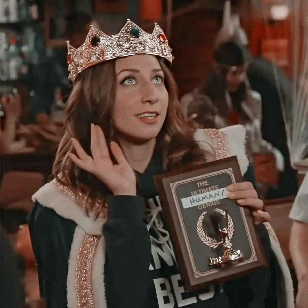 A woman wearing a jeweled crown and a robe with white fur trim holds a framed award labeled 'THE ULTIMATE HUMAN/ GENIUS'…