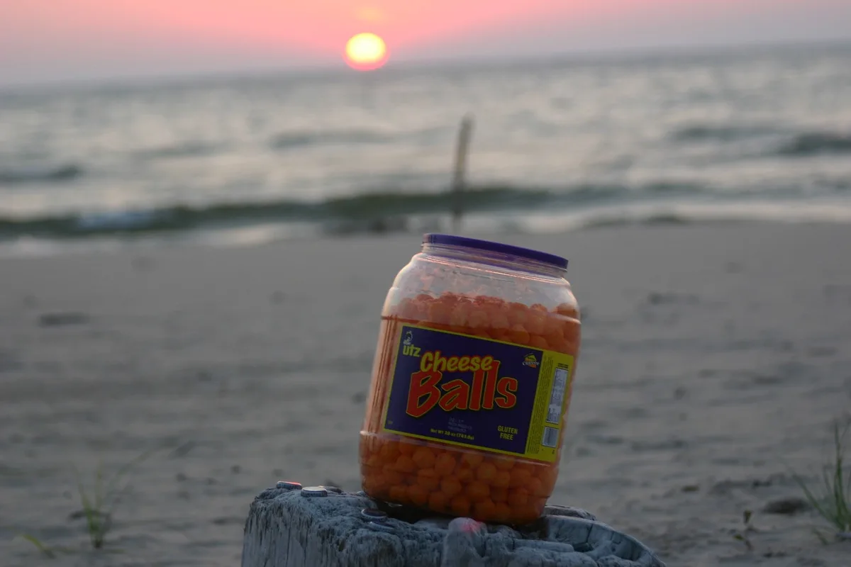 A large jar of Utz Cheese Balls sits on a weathered wooden post on a beach at sunset, with the ocean and a glowing sun in…