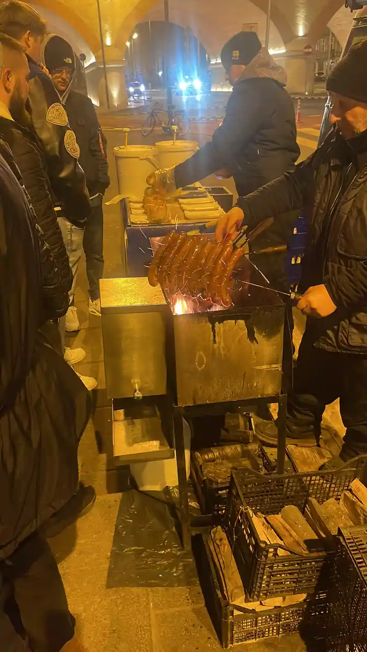 A street vendor grills numerous sausages on a metal spit over an open flame at night, while several customers in winter…