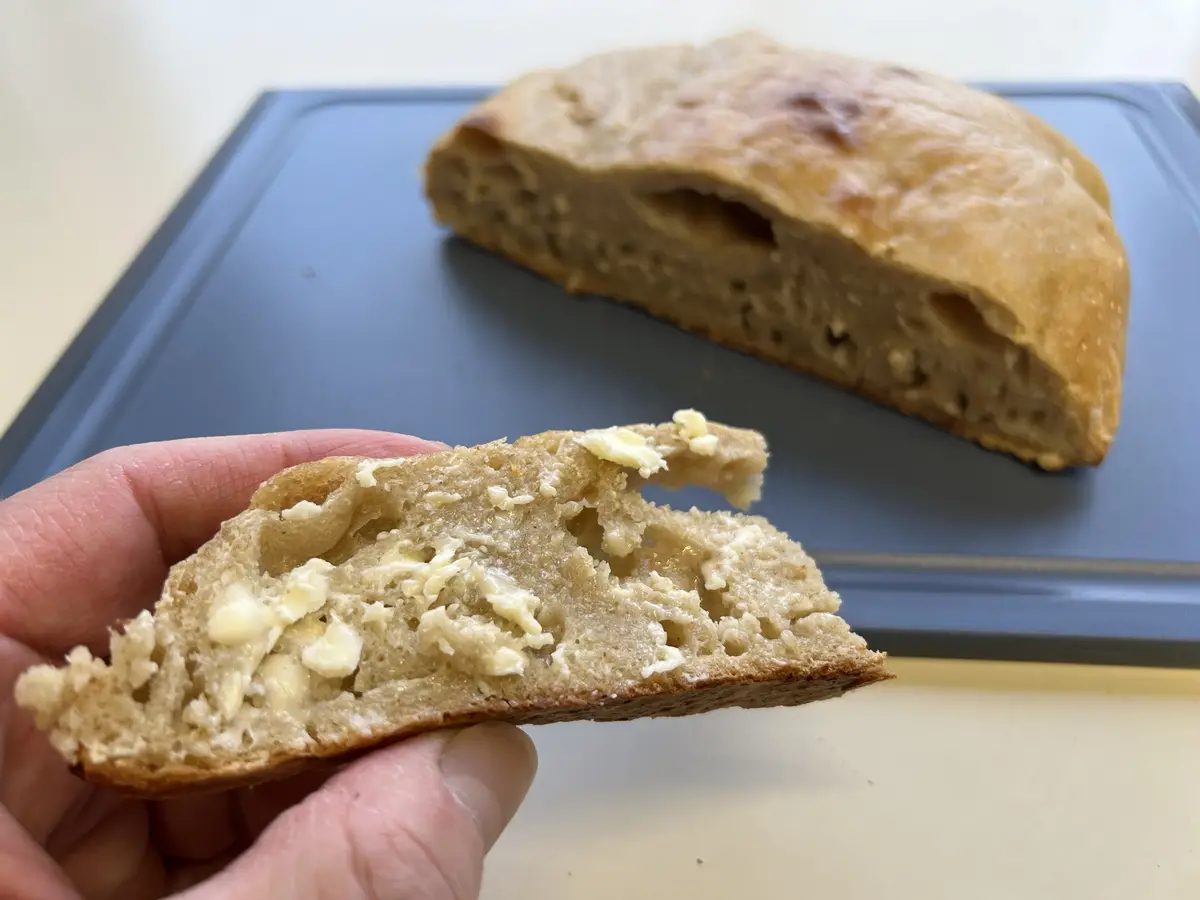 A hand holds a freshly torn slice of rustic, golden-brown bread with visible butter chunks inside, while the rest of the…