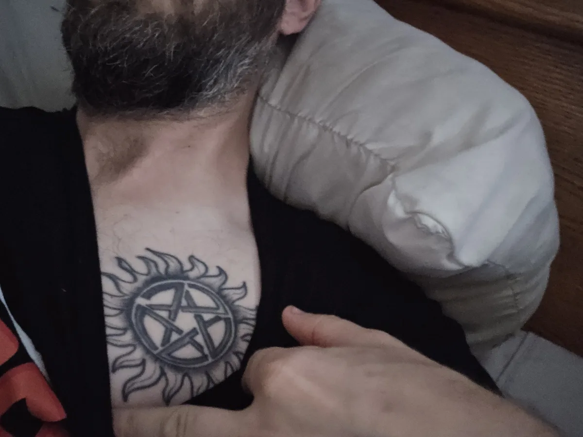 A man with a beard and black shirt is lying down, showing a black and grey tattoo on his chest that combines a pentagram…