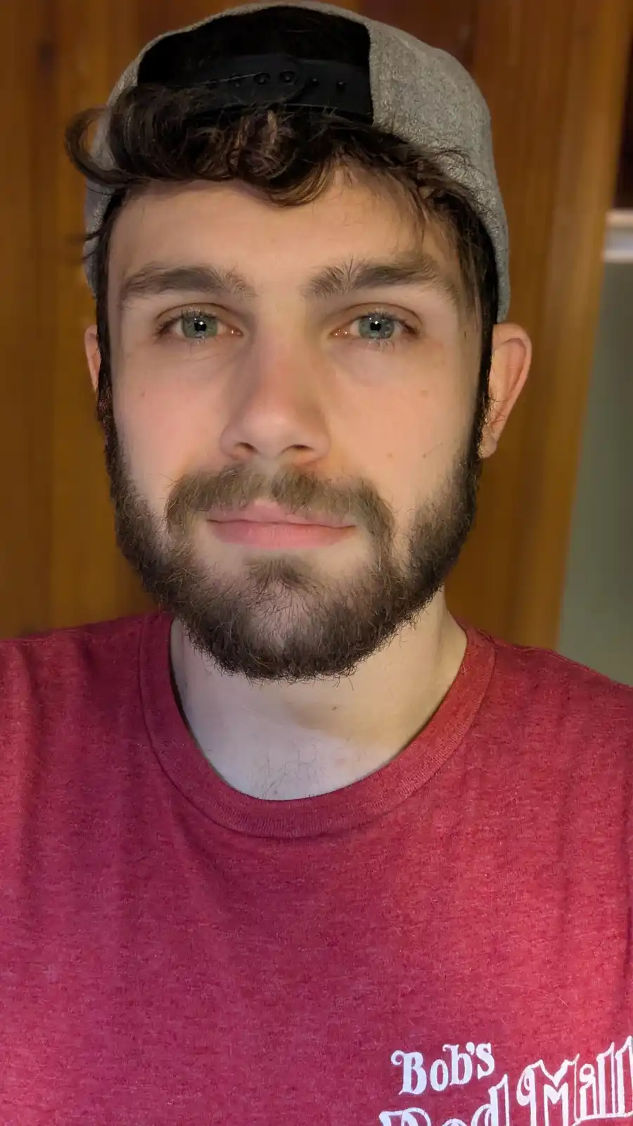 A close-up photo of a young man with curly brown hair, a beard, and a mustache, wearing a grey baseball cap backwards and a…