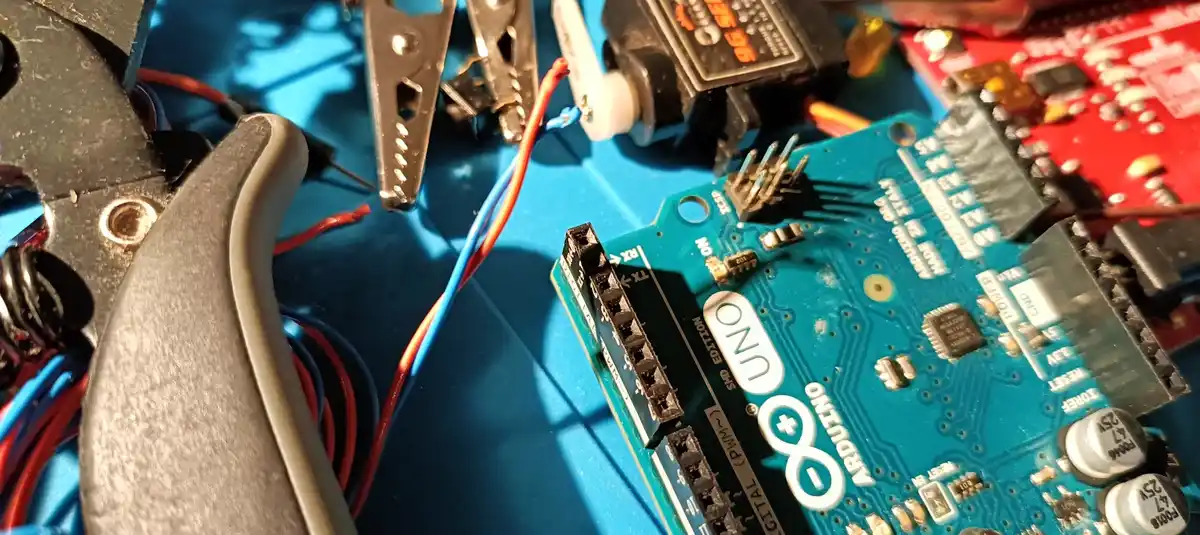 A close-up photo of an Arduino Uno microcontroller board on a blue work surface, surrounded by electronics tools like…
