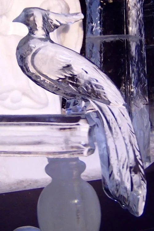 A close-up photograph of a detailed, transparent ice sculpture of a bird perched on a decorative ice base. The bird has a…