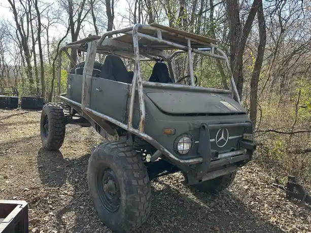 A heavily modified, olive-green Mercedes-Benz Unimog-style off-road vehicle with a metal roll cage, large knobby tires, and…