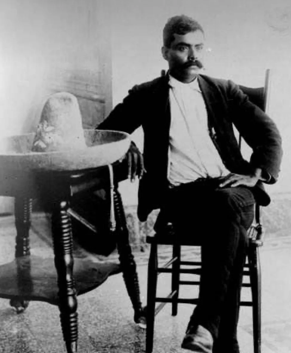 a black-and-white historical photograph shows a man with a mustache sitting in a chair, looking directly at the camera…