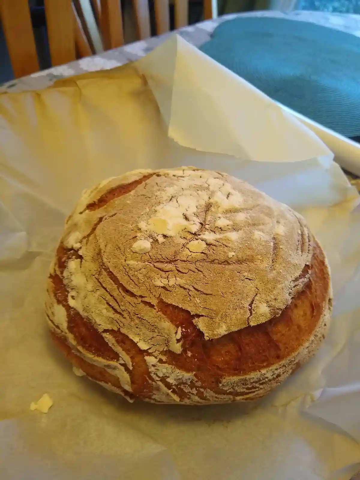 A round, rustic loaf of bread with a cracked, flour-dusted crust sits on parchment paper on a dining table. The bread has a…