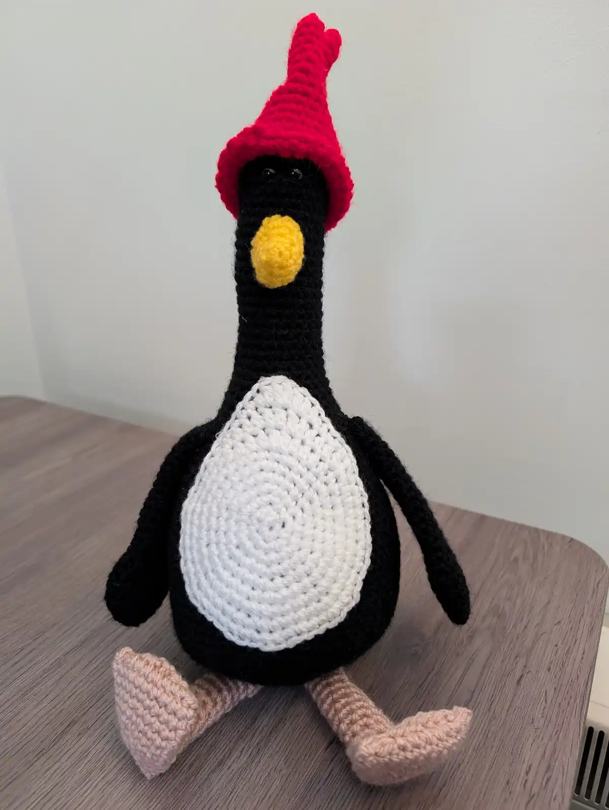 A handmade crocheted penguin plushie with a black body, white belly, yellow beak, and pink feet sits on a wooden table. It…
