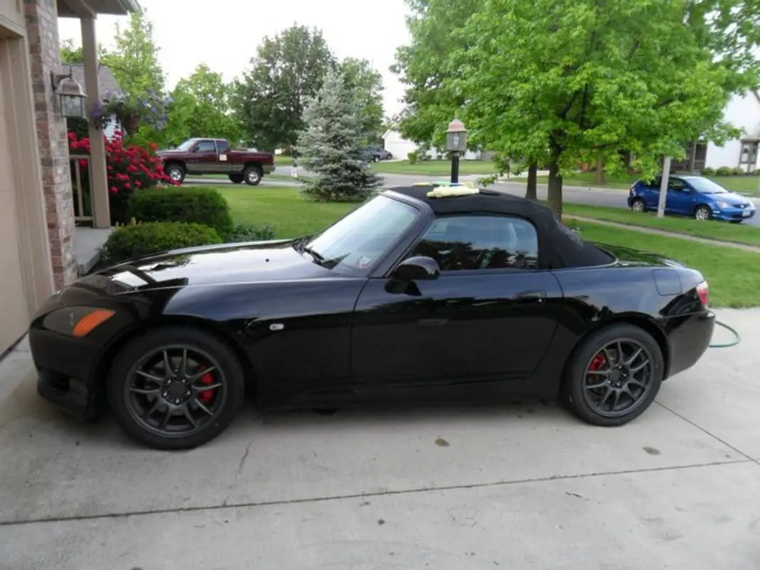 A black Honda S2000 convertible is parked on a concrete driveway in a suburban neighborhood, with trees and other cars…