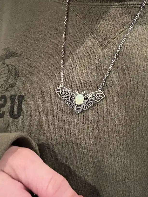 A close-up photo shows a silver-colored necklace with a delicate, filigree-style bee pendant, featuring a light-colored…