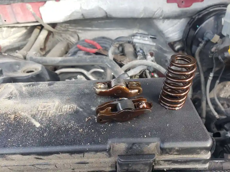 A close-up photo shows two valve rocker arms and a coil spring resting on a dusty black engine cover inside an open car…