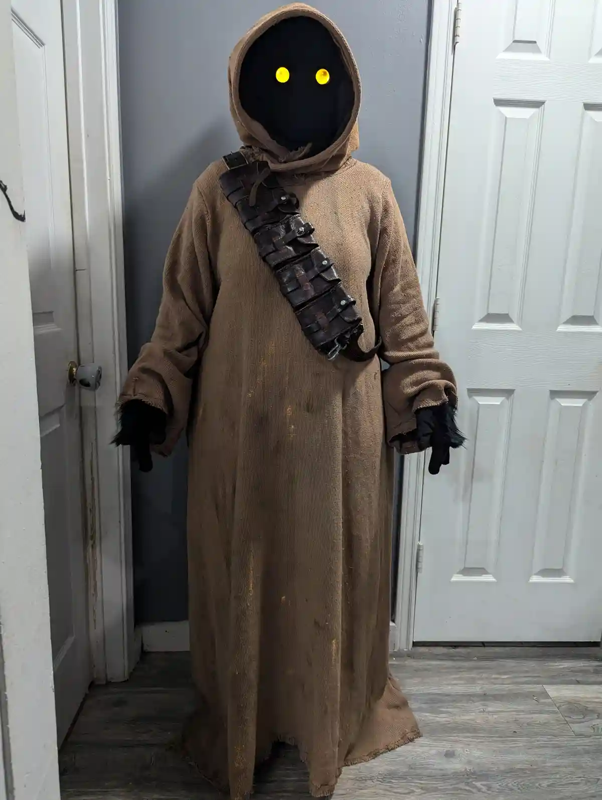 A person in a brown, hooded robe with glowing yellow eyes stands in a doorway, wearing a leather utility belt and black…