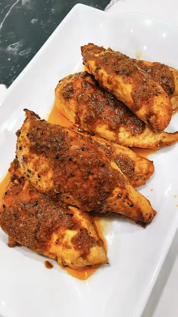 A close-up photo shows four cooked chicken breasts on a white rectangular plate, covered in a dark, glossy sauce with…