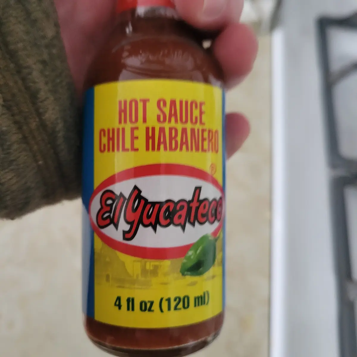 A hand holds a small glass bottle of El Yucateco brand hot sauce labeled 'Chile Habanero' with a yellow label, red text, and…