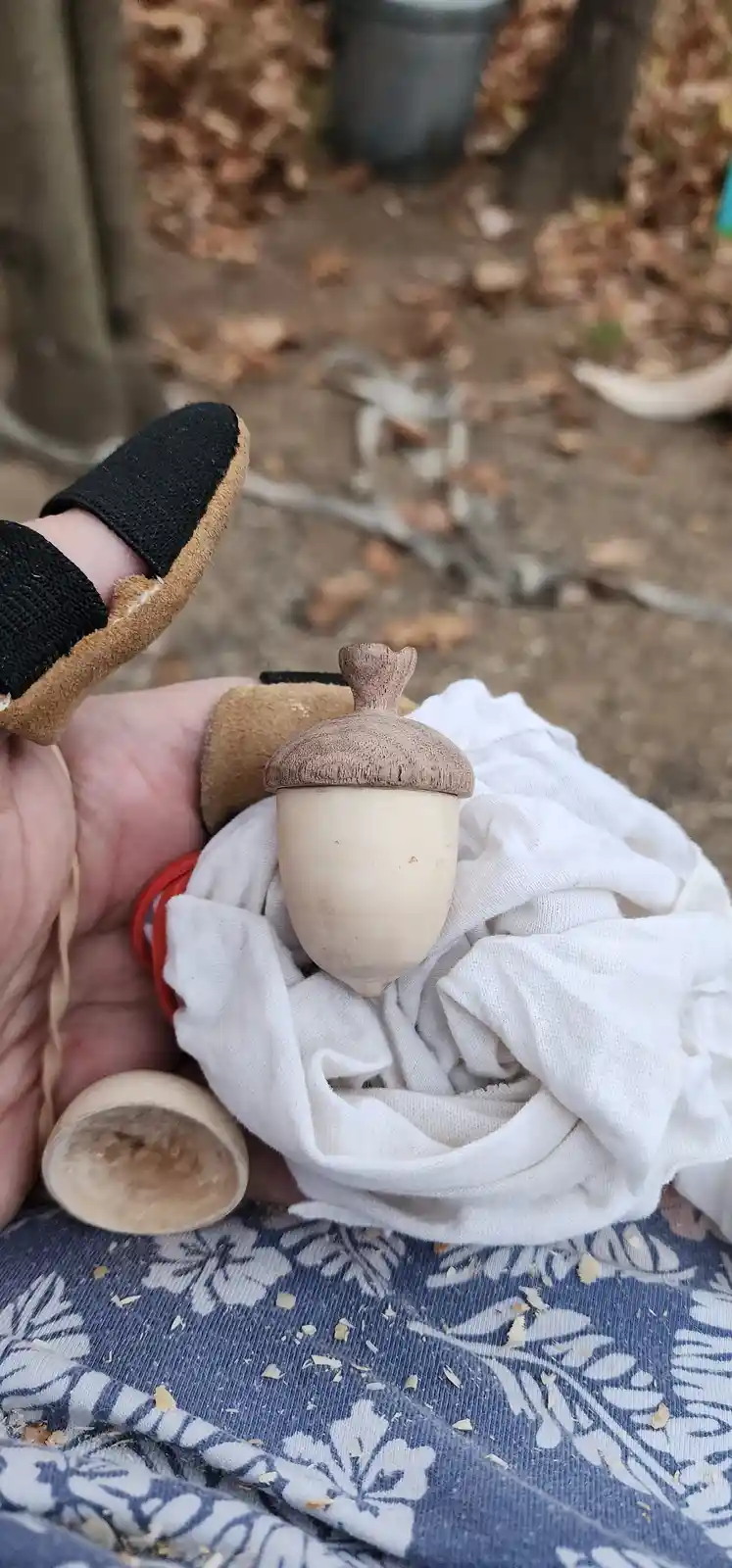 A hand wearing a black and tan protective glove holds a freshly carved wooden acorn, wrapped in a white cloth. Wood shavings…