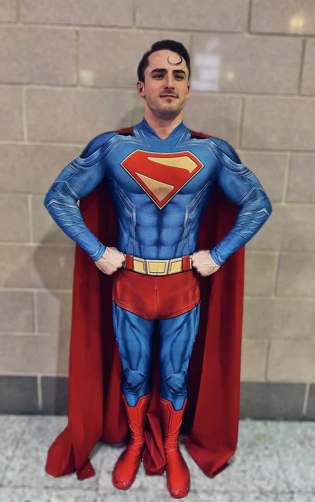 A man in a detailed Superman costume with a red cape, blue suit, and red boots stands with hands on hips against a tiled…