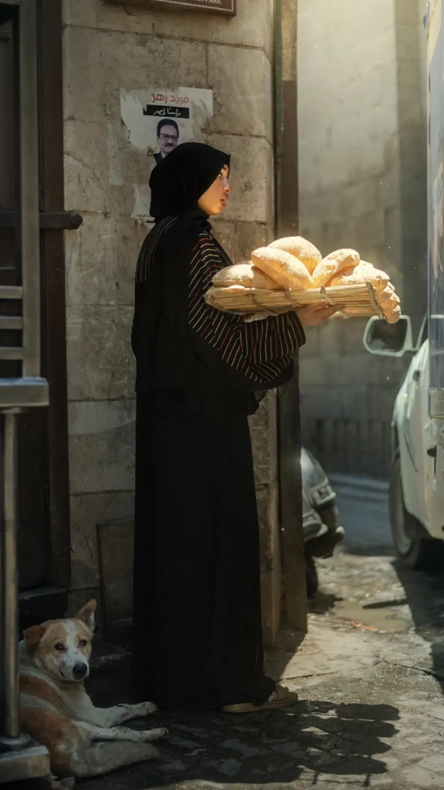 A woman in a black hijab and long black robe stands in a sunlit alleyway holding a woven tray piled high with freshly baked…