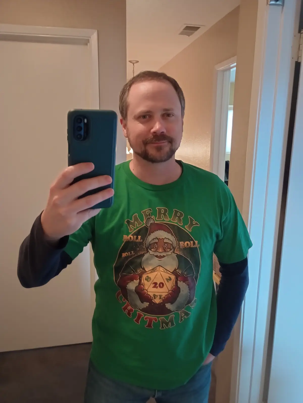 A man takes a mirror selfie holding a phone, wearing a green t-shirt with a graphic of Santa Claus holding a 20-sided die…