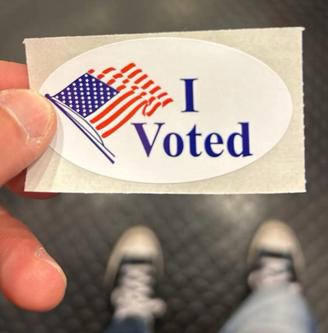 A close-up photo shows a person holding an oval 'I Voted' sticker featuring a stylized American flag, with their feet and…