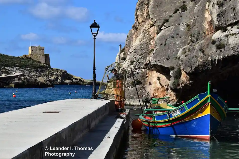 A fisherman in orange overalls stands on a concrete pier next to a colorful blue, green, and yellow boat, holding a large…