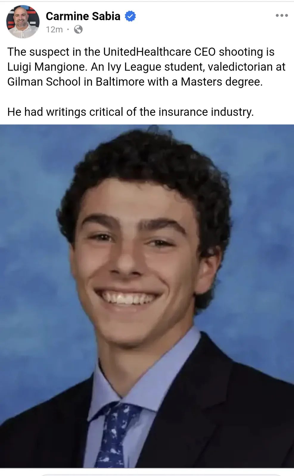 A young man with curly dark hair, wearing a dark suit, light blue shirt, and patterned tie, smiles broadly in a formal…