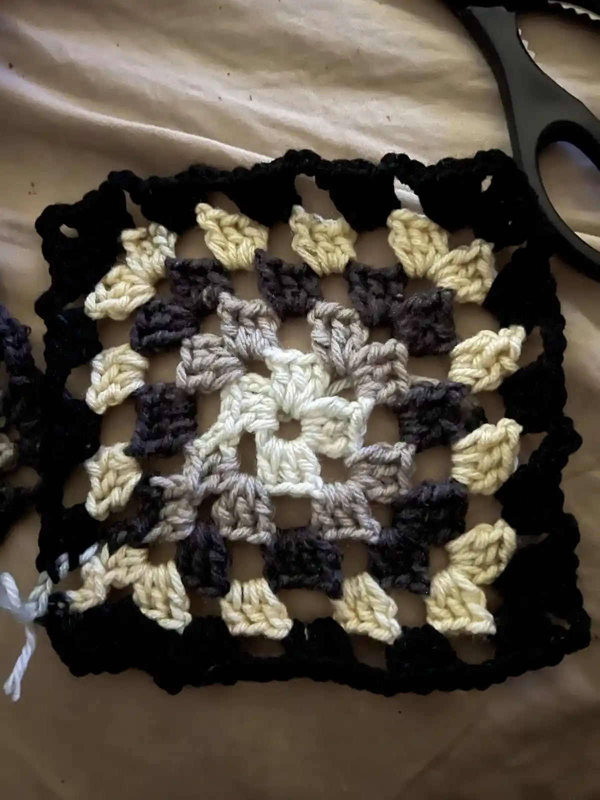A close-up photo shows a handmade crocheted square with a black border, concentric rings of cream, gray, and dark gray yarn…