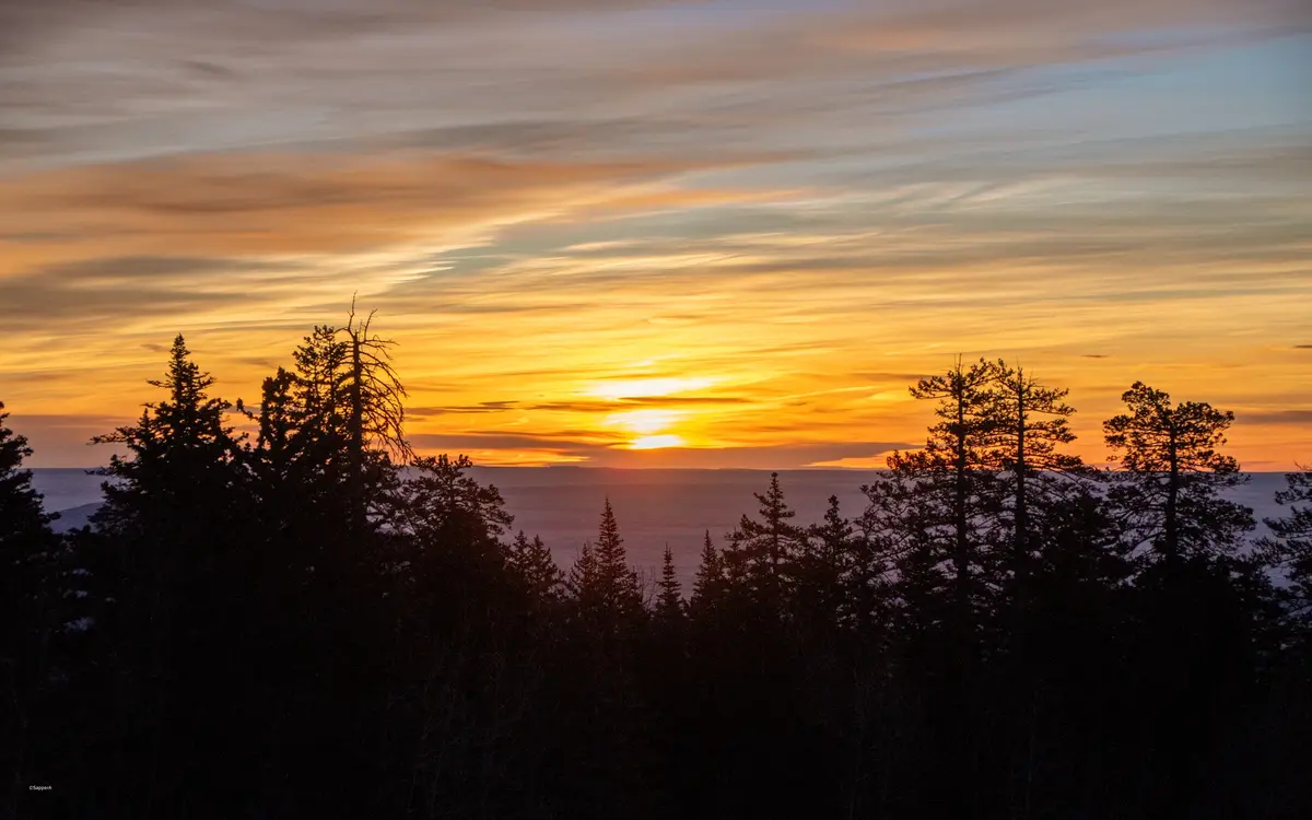 A serene landscape photo of a vibrant orange and yellow sunset over distant mountains, framed by the dark silhouettes of…