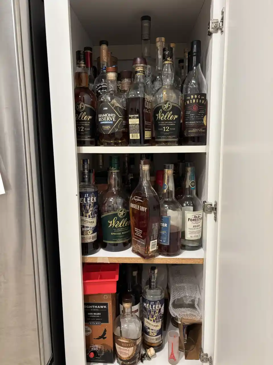 A white cabinet door is open, revealing three shelves packed with various bottles of whiskey and bourbon, including Weller…