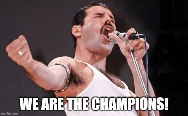 A man with dark hair and a mustache, wearing a white tank top and a chain bracelet, sings passionately into a microphone…