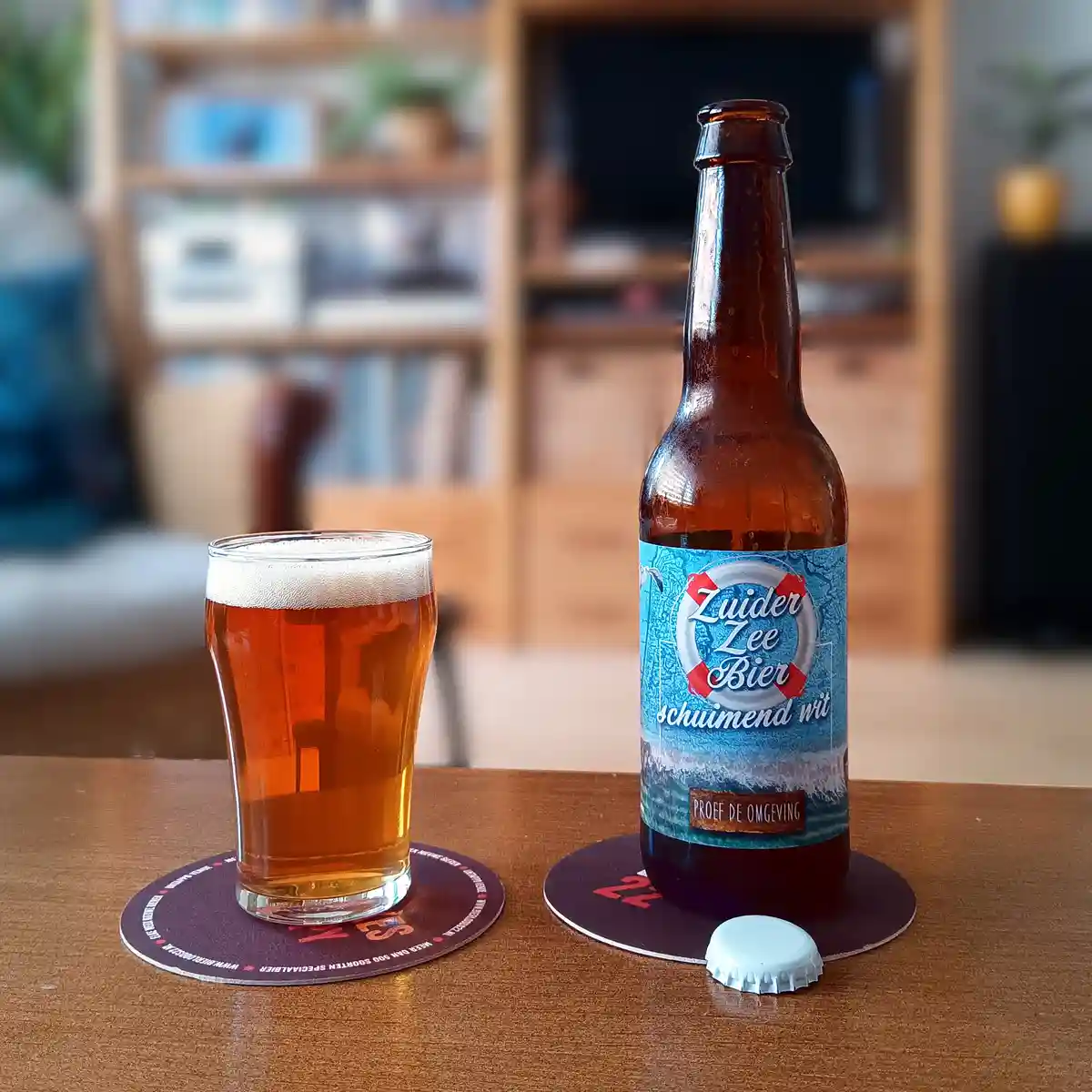 A brown beer bottle with a blue label reading 'Zuider Zee Bier schuimend wit' and 'PROEF DE OMGEVING' sits next to a glass…