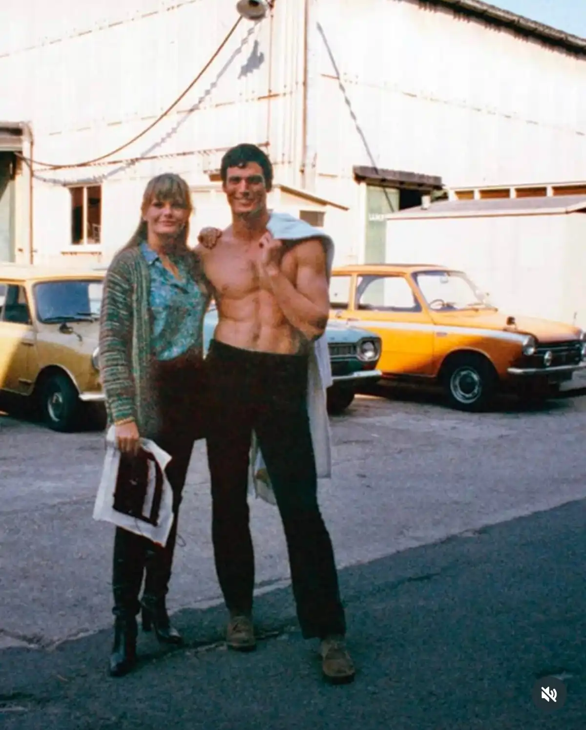 A vintage photo shows a muscular, shirtless man posing with a woman in front of a white building with two vintage cars…