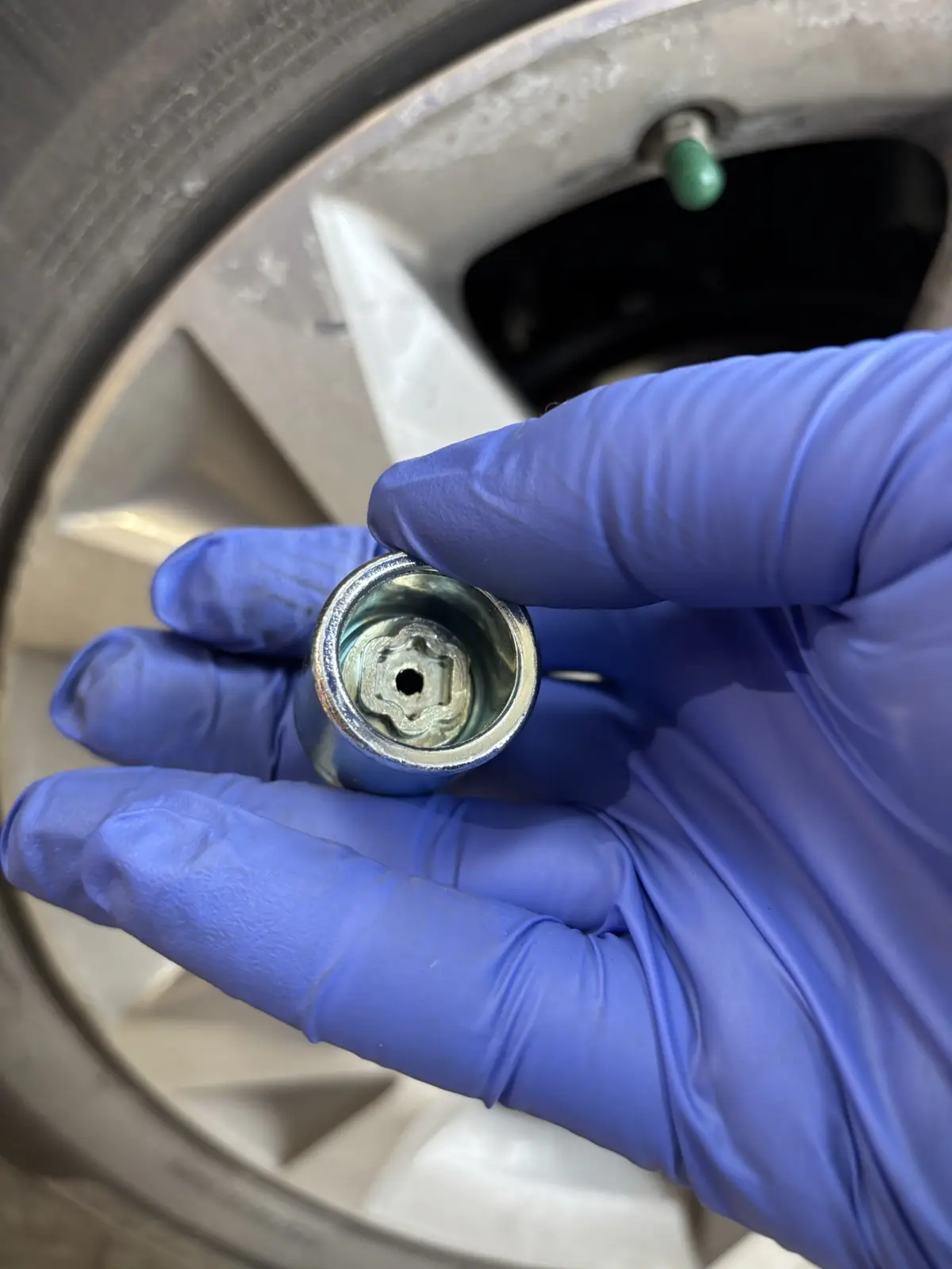 A hand wearing a blue nitrile glove holds a socket wrench with a star-shaped socket, positioned in front of a car wheel with…