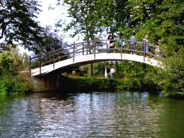 A white arched pedestrian bridge spans a calm river, surrounded by lush green trees and bushes. Several people are visible…