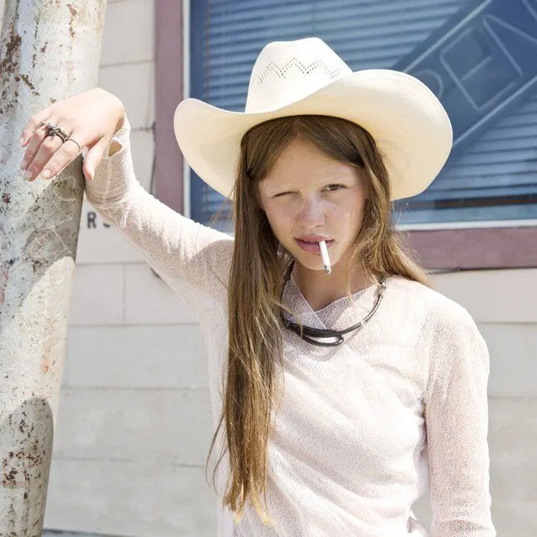 a young girl with long brown hair wears a white cowboy hat and a light-colored long-sleeved shirt, leaning against a tree…