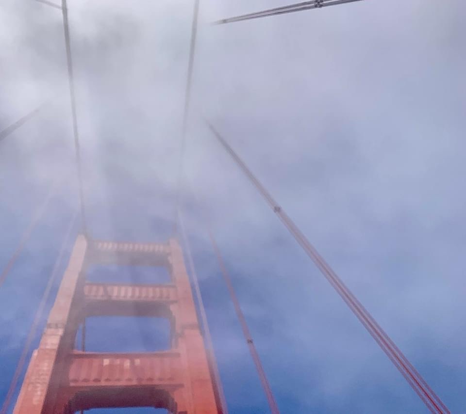 A low-angle photo of the Golden Gate Bridge partially obscured by thick fog, with its orange-red tower and suspension cables…