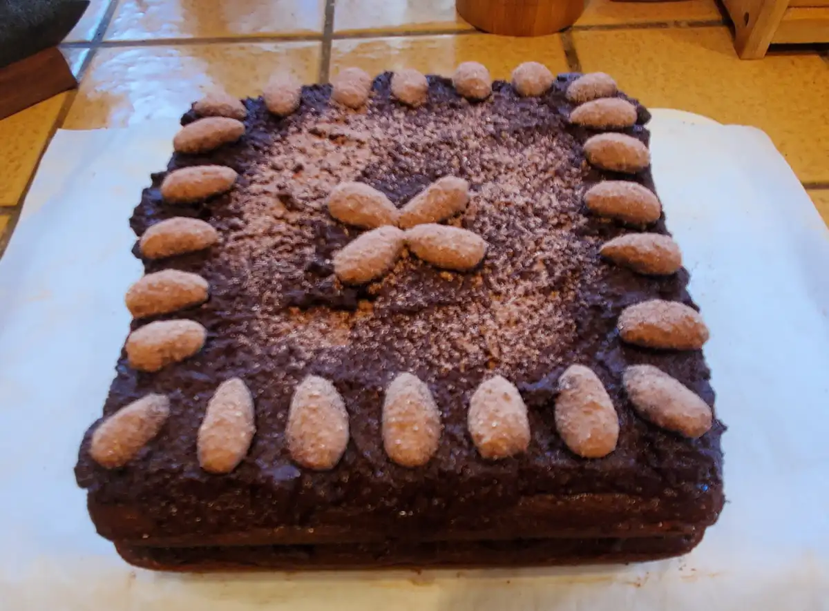 A rectangular chocolate cake with chocolate frosting, decorated with a border of sugared almonds and a central almond cross…