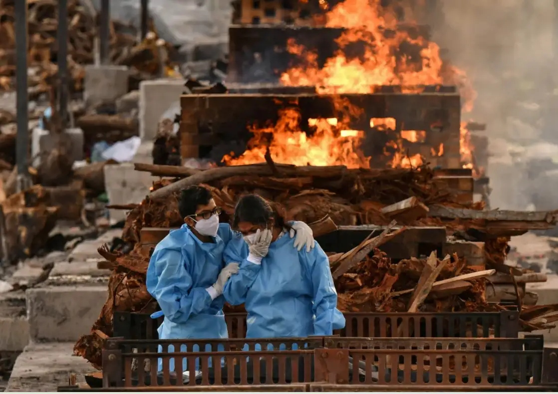 Two people in blue protective suits and masks stand near a large fire burning wooden debris; one person comforts the other…