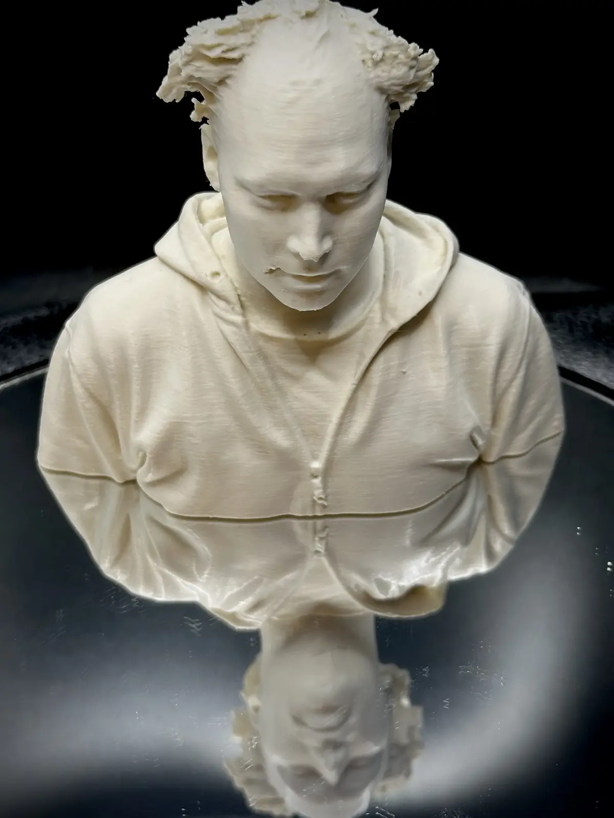 A white 3d-printed bust of a person wearing a hoodie, with visible layer lines and a slightly messy hairline, sits on a…