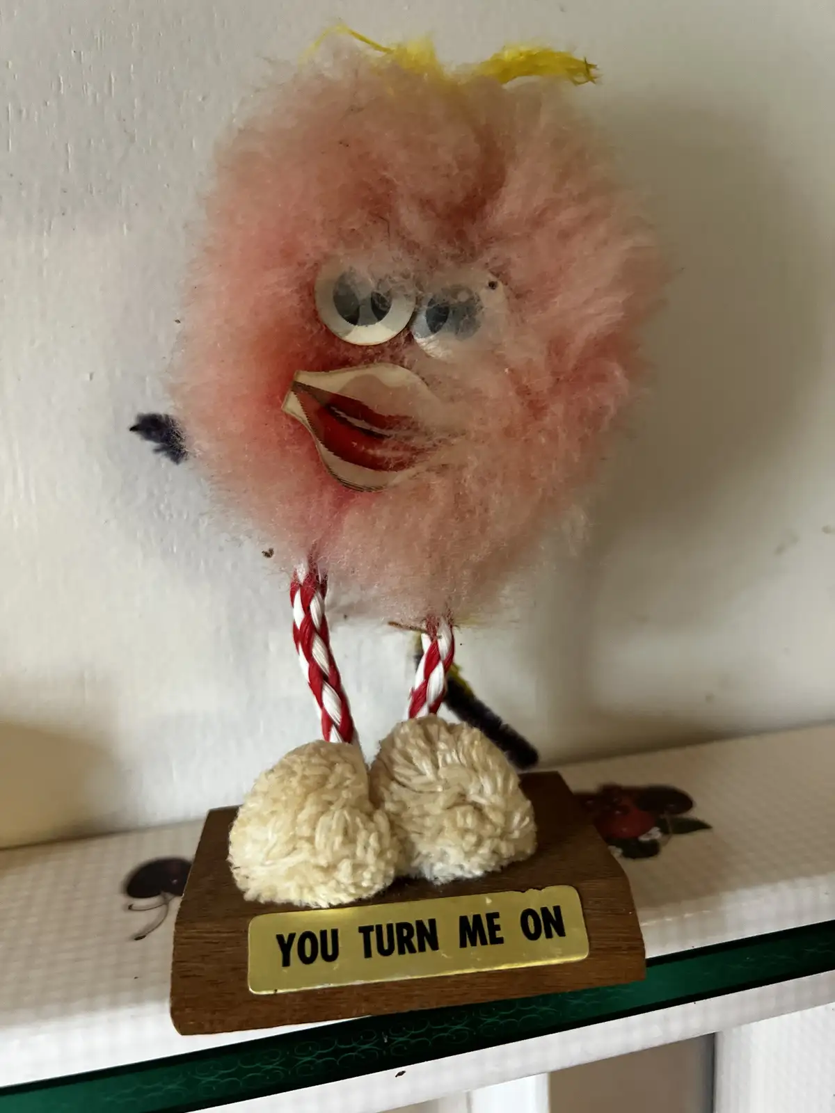 A handmade pink fuzzy bird figurine with googly eyes, red-and-white striped legs, and yarn feet stands on a wooden base with…