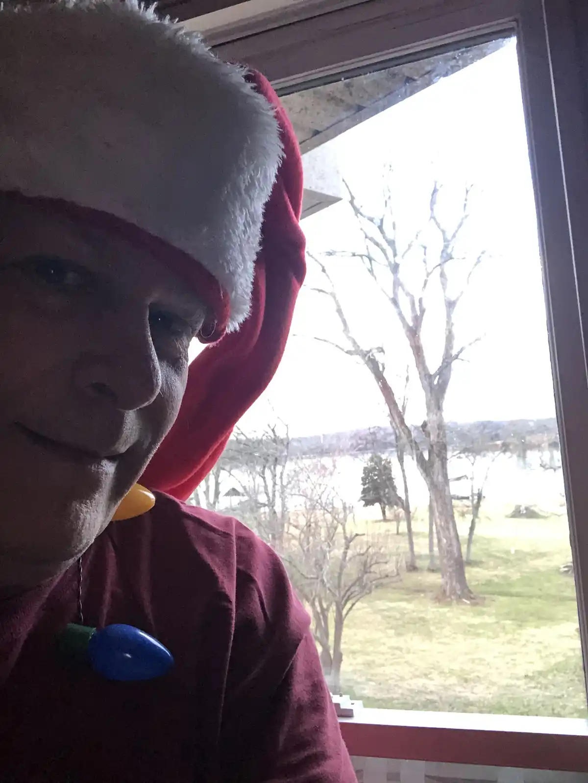 A man wearing a red and white Santa hat and a maroon shirt with a blue and yellow Christmas light bulb pin looks at the…
