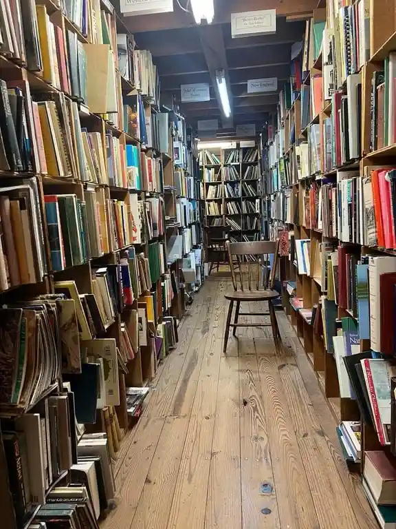 A single wooden chair sits centered in a narrow aisle of a bookstore, flanked by towering, densely packed bookshelves that…