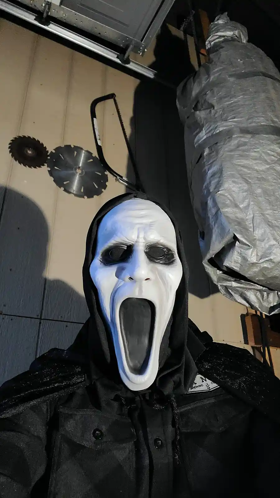 A person wearing a white Ghostface mask from the Scream movies, with a black hooded jacket, has two circular saw blades…