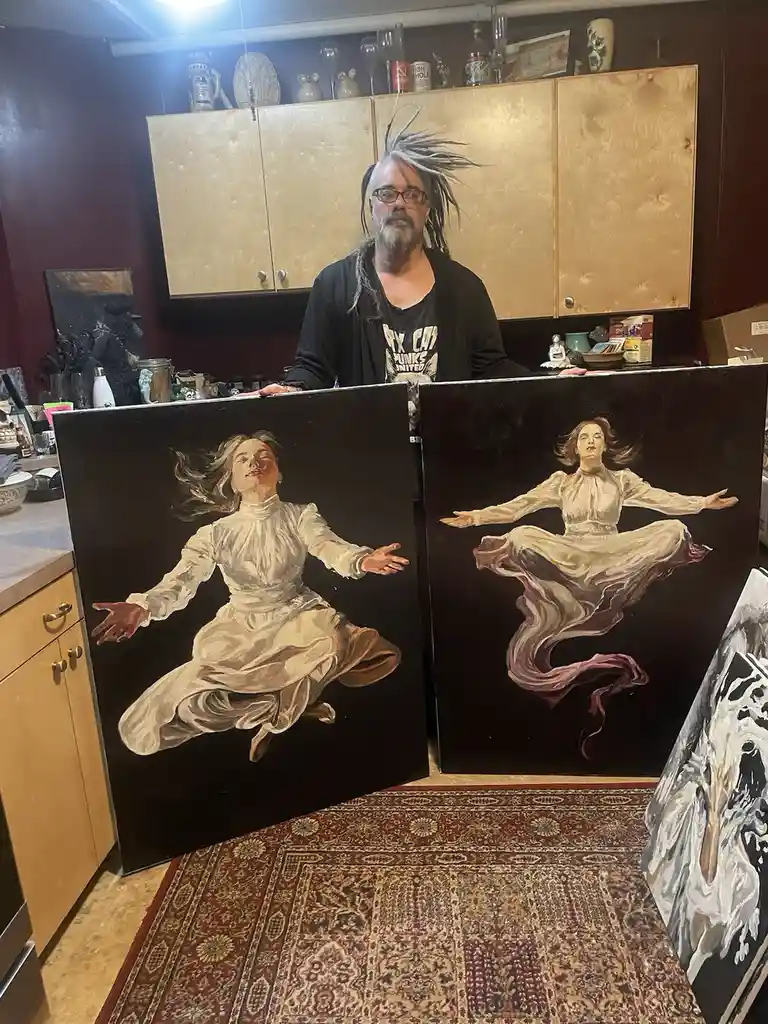 A man with dreadlocks and glasses stands in a kitchen holding two large paintings of a woman in flowing white dresses, both…