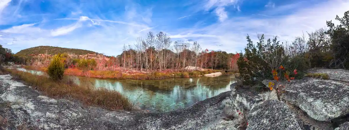 A panoramic photo of a serene river surrounded by autumn foliage, with red and orange leaves on trees and bushes, and large…