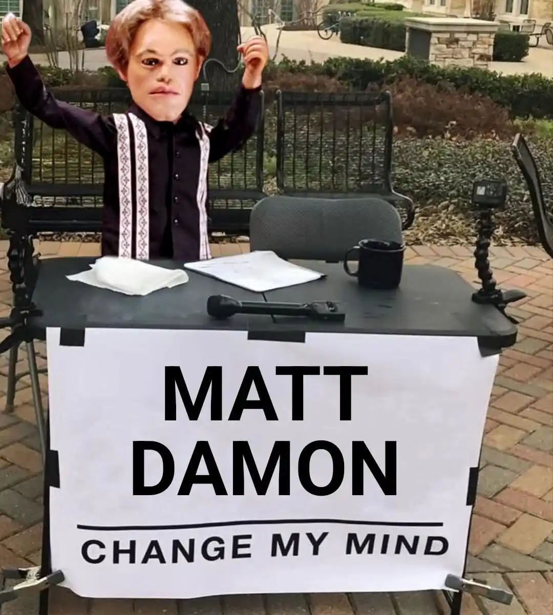 A person wearing a large Matt Damon mask sits at a makeshift outdoor desk with a microphone, papers, and a mug, arms raised…