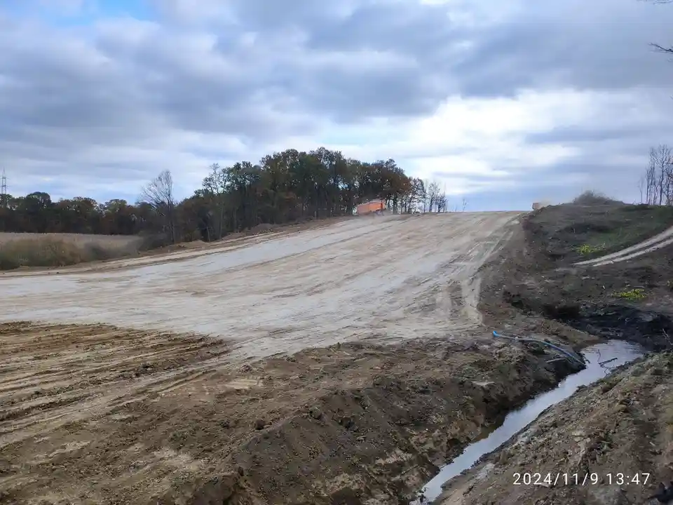 A wide, muddy construction site with a steep, freshly graded dirt hill under a cloudy sky. An orange dump truck is visible…