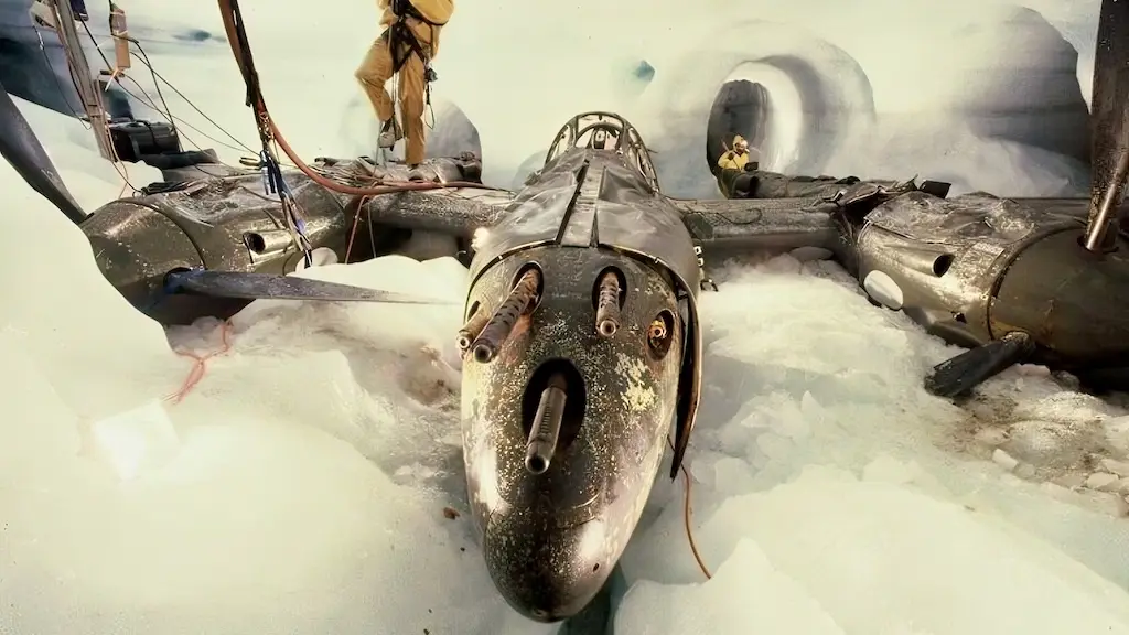 a damaged, dark-colored military aircraft lies embedded in thick white snow and ice, with two people in tan winter gear…