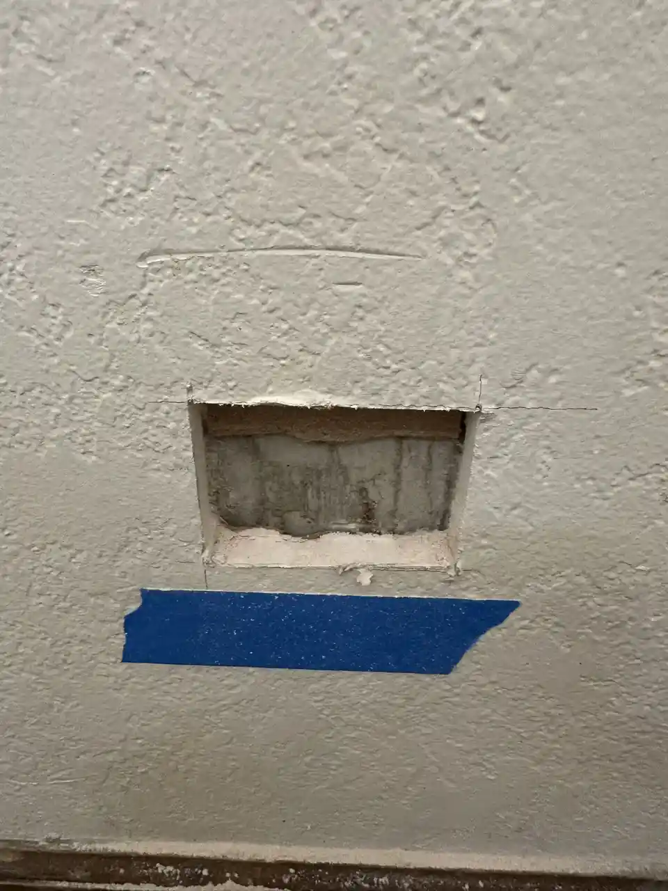 A close-up photo shows a roughly cut rectangular hole in a textured white wall, revealing a darker interior surface. Below…